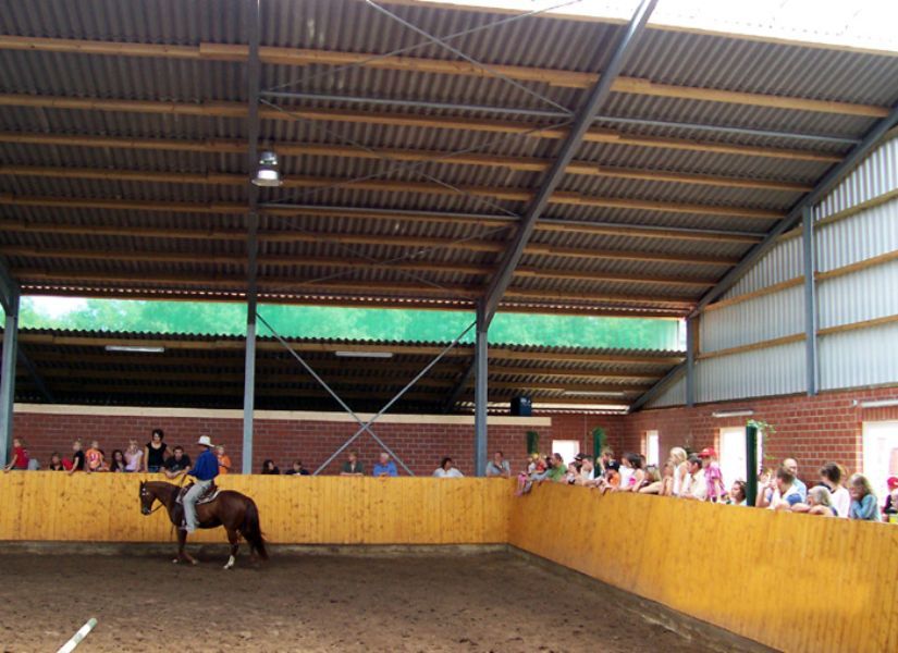 Innenbereich einer Reithalle mit Mann mit Cowboyhut auf einem Pferd und Zuschauern